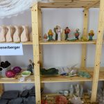 Drewniany regał z ceramicznymi figurkami i materiałami rzemieślniczymi. Kolory są stonowane, dominują beże i zielenie. Obok znajduje się kosz i białe zasłony.