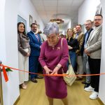Starsza kobieta w fioletowej sukience przecina czerwoną wstęgę w korytarzu. W tle kilkanaście osób. Pomieszczenie jest jasne, z białym sufitem.
