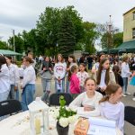 Grupa młodzieży w jasnych ubraniach stoi na zewnątrz przed budynkiem. W tle zielone namioty i drzewa, pogodny dzień. Na pierwszym planie stół z białym obrusem, świecznikiem i popcornem.