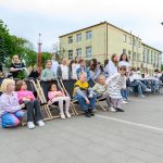 Grupa dzieci i młodzieży na plenerowym wydarzeniu w otoczeniu budynków. Kolorowe ubrania; pogodny, jasny dzień. Tło z drzewami i krzesłami.