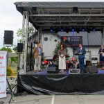 Scena z kilkoma osobami, stoisko z logo Mazowsza po lewej. Tło z szarym budynkiem, czarny dach nad sceną. Kolory głównie stonowane.