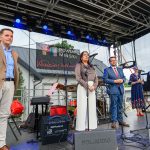 Na scenie stoi pięć osób przemawiających do mikrofonu. Tło stanowi baner z logiem i napisem Powiat Miński. Scena jest zadaszona, wokół sprzęt audio i instrumenty.
