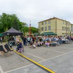 Plac przed budynkiem z tłumem ludzi. Zielone i czarne namioty. Dzieci siedzące na leżakach na szarej kostce.