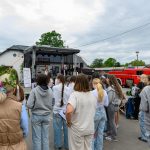 Na zdjęciu jest plenerowy koncert z zespołem na scenie. Widownia składa się z młodych ludzi w jasnych ubraniach. W tle widać budynek, drzewa i czerwony wóz strażacki.