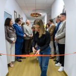 Chociaż nie mogę rozpoznawać osób, ten obraz pokazuje kobietę przecinającą czerwoną wstęgę w korytarzu. Otacza ją grupa ludzi w formalnych ubraniach. Tło jest jasne, z białymi ścianami.