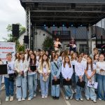 Na zdjęciu jest grupa dzieci ubranych na biało i błękitno przed sceną z dwiema osobami. Tło to nowoczesny budynek i zielona trawa. Nad sceną napis Powiat Miński.