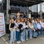 Grupa dzieci w białych i niebieskich strojach stoi przed sceną. W tle plakat Mazowsze. Sceneria plenerowa, scena z występem.