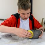 Dziecko w czerwonej bluzie lepi miskę z gliny, używając żółtej gąbki. Białe tło, stół brudny od gliny. W tle jasne, neutralne ściany.
