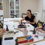 Trzy kobiety tworzą ceramiczne przedmioty przy dużym stole z narzędziami. W tle białe regały z książkami i ceramiką. Pomieszczenie jest jasne i przytulne.