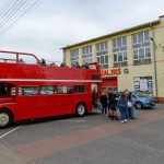 Czerwony piętrowy autobus stoi przed żółtym budynkiem z napisem OCHOTNICZA STRAŻ POŻARNA 1915. Na górnym pokładzie autobusu siedzą ludzie. Kilka osób stoi obok pojazdu.