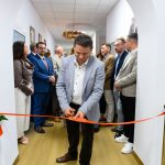Osoba w jasnej marynarce przecina czerwoną wstęgę w korytarzu. W tle stoją elegancko ubrani ludzie. Jasne ściany i drewniana podłoga.