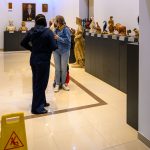 W galerii sztuki na jasnej podłodze stoją rzeźby i obrazy. Dwie osoby rozmawiają, a obok znak śliska podłoga. Ściany są białe, a rzeźby wystawione na ciemnych cokołach.