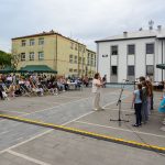 Na zdjęciu grupa dzieci na scenie, ubrana kolorowo, śpiewa dla publiczności na szarym placu z budynkami w tle.