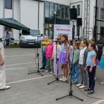 Grupa dzieci w kolorowych ubraniach śpiewa na scenie z mikrofonami. Przed nimi stoi kobieta. W tle biały budynek i samochód.