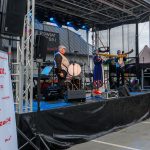 Zespół muzyczny na scenie na tle budynku, gra na instrumentach. Dominują kolory niebieski, czarny i czerwony. Baner z napisem Mazowsze.