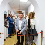 Mężczyzna przecina czerwoną wstęgę, otoczony przez ludzi w jasnym korytarzu. W tle białe ściany, drewniana podłoga. Wszyscy elegancko ubrani.