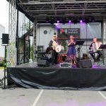 Trzech muzyków gra na scenie: kobieta śpiewa, mężczyzna gra na akordeonie, trzeci na bębnie. W tle banner Powiat Miński. Tło jasne, szaro-białe budynki.
