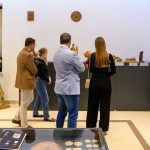 Grupa osób oglądająca wystawę rzeźb w jasnej sali muzeum. Dominują kolory beżowy i szary. W tle na ścianie wiszą małe płaskorzeźby.