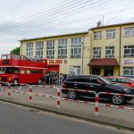 Czerwony, piętrowy autobus stoi przed żółtym budynkiem z napisem Dom Strażaka. Na ulicy kilka zaparkowanych samochodów. Grupa ludzi stoi obok autobusu.