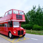 Czerwony piętrowy autobus jedzie po drodze. W tle zielone drzewa. Niebo lekko zachmurzone.