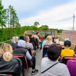 Grupa ludzi siedzi na piętrze czerwonego autobusu z otwartym dachem. Wokół zielone drzewa i jasne niebo. Obok mija ogrodzenie z cegieł.