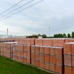 Stosy cegieł zapakowane w folię stoją na zielonej trawie. Tło to zachmurzone niebo i drzewa. Widoczny jest także budynek fabryki.