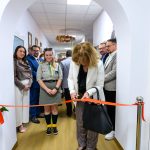 Funkcjonariuszka przecina wstęgę w jasnym korytarzu, otoczona ludźmi. Kobieta w białej marynarce i wzorzystych spodniach. Drewniana podłoga, ściany białe z zawieszonymi dekoracjami.