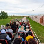 Ludzie siedzą na otwartym piętrze czerwonego autobusu i patrzą na fabrykę cegieł. Po lewej stronie zielona trawa i drzewa, po prawej stosy cegieł. Niebo jest pochmurne.