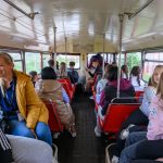 Ludzie siedzą w starym autobusie z czerwonymi siedzeniami. Tło jest zielone z widokiem przez okna. Kobieta w żółtej kurtce uśmiecha się.