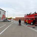Czerwony, dwupoziomowy autobus stoi na placu z ludźmi na górnym pokładzie. Szary budynek po lewej i stosy cegieł w tle. Pochmurny dzień.