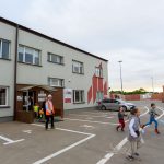 Na zdjęciu znajduje się szara, dwupiętrowa budynek z czerwonymi elementami. Przed nim ludzie, w tym dzieci biegające po parkingu. W tle widać stosy cegieł i fragmenty zieleni.