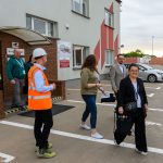 Na zdjęciu widać grupę ludzi na placu fabrycznym. Budynek z czerwoną i szarą fasadą oraz stosy palet w tle. Jeden z mężczyzn nosi pomarańczową kamizelkę odblaskową.