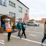 Grupa ludzi wychodzi z budynku w beżowo-czerwonym kolorze. Mężczyzna w pomarańczowej kamizelce stoi na zewnątrz. W tle, parking i stosy cegieł.