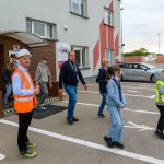 Grupa ludzi w kamizelkach odblaskowych wychodzi z budynku z cegły. Na parkingu widać samochód i stosy kartonów. Tło jest zachmurzone.