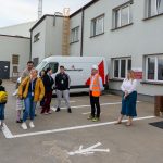 Grupa osób przed zakładem przemysłowym, z białym vanem w tle. Jasne budynki, niektóre osoby w kamizelkach odblaskowych. Tło jest szare i przemysłowe.