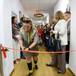 Osoba w harcerskim mundurze przecina czerwoną wstęgę w jasnym korytarzu. Tło: biały sufit, grupa ludzi. Na ziemi jasne, drewniane panele.