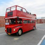 Czerwony piętrowy autobus jedzie po betonowej powierzchni placu. W tle stosy kartonowych pudeł i budynek. Na górnym pokładzie siedzą pasażerowie.