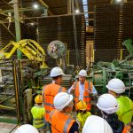 Grupa osób w hełmach i pomarańczowych kamizelkach jest w fabryce. Tło to maszyny i duże metalowe stosy. Przewodnik wyjaśnia procesy produkcyjne.