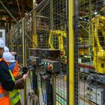 Pracownicy w kaskach i kamizelkach obserwują żółte roboty przemysłowe przez metalową kratę. Tło to hala fabryczna z metalowymi konstrukcjami. Dominują kolory: żółty, pomarańczowy i metaliczny.