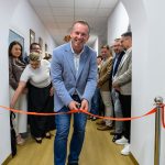 Mężczyzna w niebieskiej marynarce przecina czerwoną wstęgę podczas ceremonii otwarcia. W tle grupa uśmiechniętych osób na korytarzu. Dominują kolory bieli i drewna.