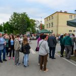 Grupa ludzi na placu przed budynkami, zielone i beżowe namioty. Drzewa w tle, niebo jasne, ludzie rozmawiają.