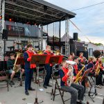 Orkiestra na scenie, młodzież w granatowych uniformach z czerwonymi elementami. Tło stanowi namiot z napisem Powiat Miński. Obok scena z instrumentami i mikrofonami.