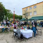 Na zdjęciu widać grupę ludzi na pikniku, siedzących przy stołach na zewnątrz. W tle jest zielony namiot z napisem oraz żółty budynek. Trawa i drzewa otaczają miejsce wydarzenia, tworząc przyjemną atmosferę.