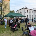 Grupa osób zgromadzona pod zielonym namiotem na tle budynku. Lato, na trawie stoliki z białymi obrusami. Swobodna, społeczna atmosfera.
