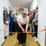 Kobieta przecina czerwoną wstęgę w korytarzu budynku. Tło jest białe z drewnianą podłogą. Obok stoi grupa elegancko ubranych osób.