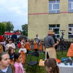 Grupa dzieci i dorosłych w fartuchach na warsztatach na świeżym powietrzu przy budynku. Stoły z kolorowymi naczyniami. W tle widać zielone drzewa i czerwony pojazd.