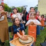 Grupa dzieci i dorośli lepioną glinę na świeżym powietrzu. Noszą fartuchy na tle zieleni i żółtej ściany. W tle widać drewniany płot i drzewa.