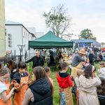 Na zdjęciu widać plenerowy festyn z zielonym namiotem, grupą dzieci w fartuszkach i dorosłych. Tło stanowią budynki i scena z oświetleniem. Atmosfera jest radosna, a trawa zielona.