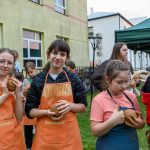 Grupa dzieci w pomarańczowych fartuchach trzymają glinę. Tło to zielony trawnik i jasnozielony budynek z oknami. W tle namiot i kilku dorosłych.