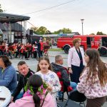 Na scenie orkiestra w czerwonych strojach przy instrumencie, w tle czerwony wóz strażacki. Przed sceną grupa ludzi siedzi i stoi, rozmawiając. Jest pogodny dzień, niebo lekko zachmurzone.